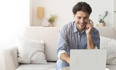 Ein junger Mann sitzt an einem Laptop. Er ist im Wohnzimmer. Er lächelt.  | © Adobe Stock